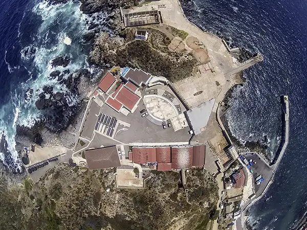 Aerial view of a rocky coastal site with red-roofed buildings around a circular central pavilion and small harbor.