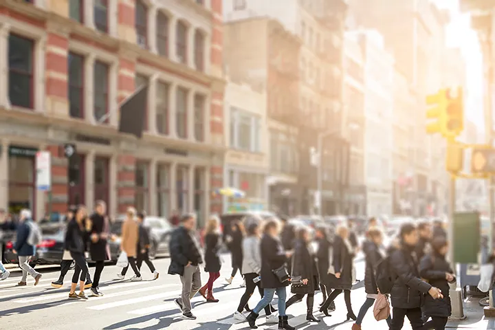 Group of pedestrians crossing a city street on a sunny day, with brick buildings and a traffic light in the background.