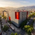 A large grey office building with red vertical banners on a city street, surrounded by trees, with mountains in the distance.