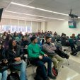 University classroom filled with students seated in rows, listening to a lecture; some take notes, others use laptops or tablets.