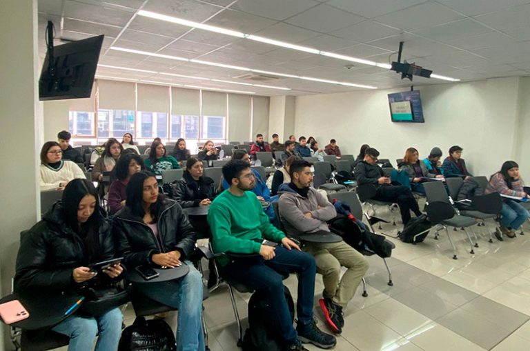 University classroom filled with students seated in rows, listening to a lecture; some take notes, others use laptops or tablets.