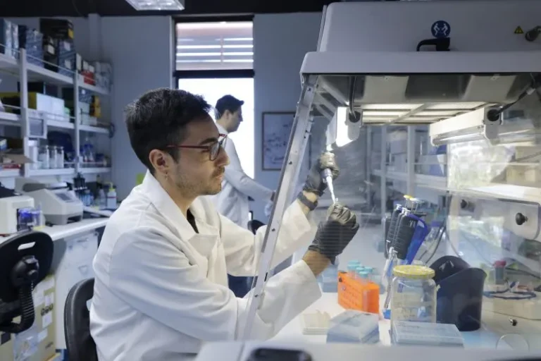 Scientist in a white lab coat uses a micropipette inside a biosafety cabinet, with lab supplies on the bench and another researcher in the background.