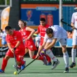 Field hockey players in red and white chase the ball on a green turf during a match.