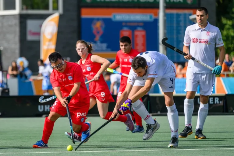 Field hockey players in red and white chase the ball on a green turf during a match.