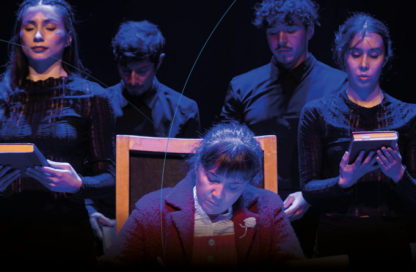 Five performers on a dark stage; three stand behind a seated woman reading a book, all holding books under blue lighting.
