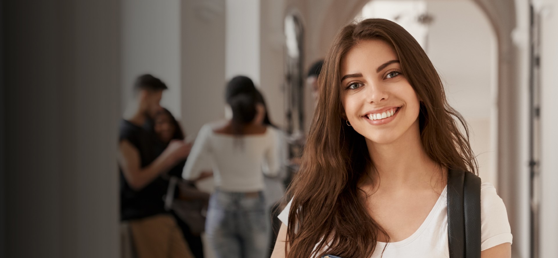 Young woman with long brown hair and a backpack smiles at the camera in a bright hallway with blurred people in the background.